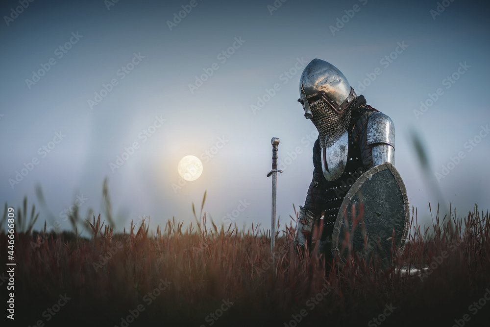 The tired knight in the plate armor kneels among the battlefield with a sword in the moon light. Lost battle. Defeat concept. - obrazy, fototapety, plakaty 