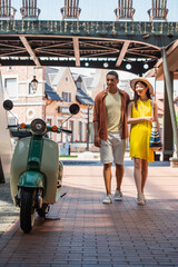 Wall Mural - stylish multiethnic couple looking at scooter while walking on urban street