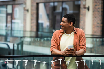 Wall Mural - smiling african american man with smartphone looking away on bridge in city