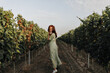 © Look! - Cheerful young girl with red hair and cool hat in trendy long dress and white sneakers smiling, looking into camera and posing on background of vineyards