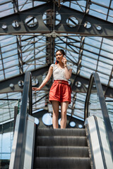 Wall Mural - low angle view of stylish asian woman talking on cellphone on escalator