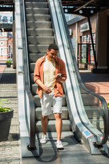 Wall Mural - cheerful african american man in shorts using mobile phone near escalator