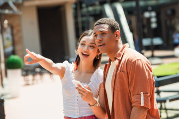 Wall Mural - amazed asian woman with smartphone pointing with finger near african american man