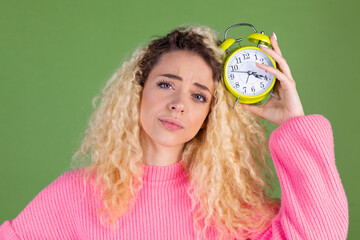 Young blonde woman with long curly hair in pink sweater on green background with alarm clock sleepy tired