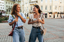 Cheerful Girls In Stylish Summer Outfits Talk And Smile. Young Attractive Blonde And Brunette Women In Jeans And Floral Blouses Walk Outside.
