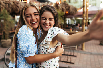 Wall Mural - Joyful brunette and blonde women hug and sincerely smile outside. Beautiful lady in white floral blouse takes selfie with girlfriend at street.