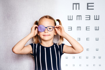 happy little girl wearing glasses and eye patch or occluder with blurry eye test chart in the backgr