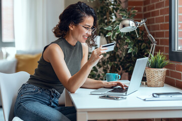 Cute woman holding white credit card for shopping online with computer while sitting in living room at home.