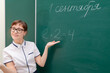 © ALEKSEI - school teacher young woman in a white blouse in the classroom on a green blackboard writes with chalk. translation - September 1. portrait