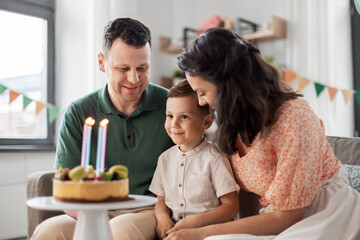 Wall Mural - family, holidays and people concept - portrait of happy mother, father and little son with four candles on birthday cake sitting on sofa at home party