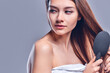 © Olena Apetroaiei - A beautiful brown-haired woman with bared shoulders looks at the camera as she brushes her hair with a brush, isolated on a gray background with copy space for text.