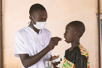 Young black medical trainee with face mask administering a tablet to a cute and compliant African boy during a healthcare campaign