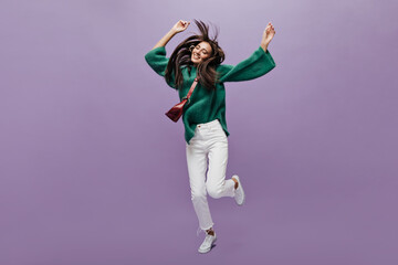 Wall Mural - Thin tanned woman in white pants and green woolen sweater dances on purple background. Charming girl with cross-body bag jumps and moves on isolated.