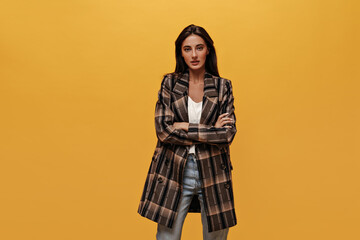 Wall Mural - Tanned brunette woman in denim pants, stylish jacket crosses arms and poses on isolated. Cool girl looks into camera on yellow background.