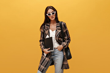 Wall Mural - Pretty woman in jeans, stylish oversized jacket and white top looks into camera. Attractive brunette girl in sunglasses holds notebooks on isolated yellow background.