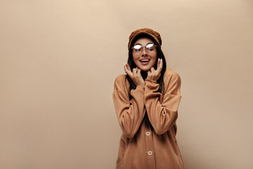 Wall Mural - Young brunette woman in eyeglasses crosses fingers. Charming girl in cap and cashmere suit smiles on isolated beige background.