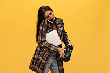 Wall Mural - Brunette long-haired woman holds black handbag. Dissatisfied girl in jeans and oversized jacket talks on phone and poses with notebooks on isolated yellow background.