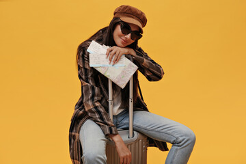 Wall Mural - Cool girl in sunglasses, beret and stylish coat smiles softly and sits on suitcase on isolated. Brunette woman in denim pants poses and holds map on yellow background.