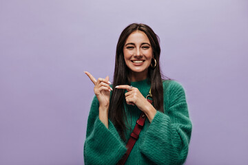 Wall Mural - Cheerful brunette woman in green stylish woolen sweater smiles sincerely. Attractive girl with cross-body bag points at place for text on purple background