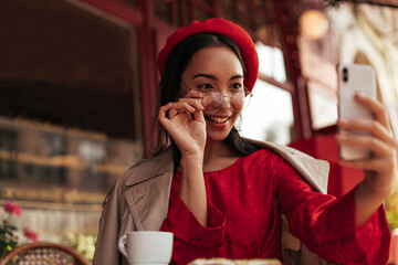 Wall Mural - Attractive brunette woman in trendy beret, red dress and beige trench coat puts off eyeglasses, holds phone and takes selfie in street cafe.