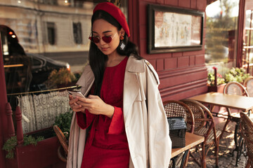 Wall Mural - Pretty brunette woman in red dress, stylish sunglasses, beret and beige trench coat holds cellphone and leans on table in street cafe.