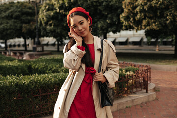 Wall Mural - Pretty young brunette woman in red dress, bright beret and beige trench coat touches face, smiles, holds handbag and poses outside.