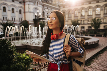 Wall Mural - Attractive brunette woman in eyeglasses, striped shirt and red silk skirt poses with headphones, holds map and backpack, smiles outside.