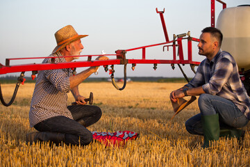 Poster - Senior farmer using wrench to fix boom sprayer and talking to young farmer.