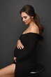 A gorgeous pregnant model girl wearing a black evening dress posing over grey background. Young pregnant woman in fashionable dress