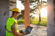 © Ольга Симонова - Construction engineer on the construction site of a house made of porous concrete blocks works at a computer in a reflective safety vest and hardhat. Design, construction, drawings project, check