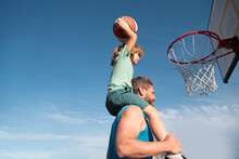 Father And Son Playing Basketball. Sports Concept. Boy Child Sitting On The Dad Shoulders, Throwing Basketball Ball Into Basket, Side View On Sky Background With Copy Space.