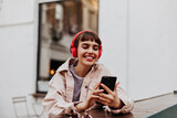 Fototapeta  - Positive short-haired girl in red headphones smiles outdoors. Brunette woman in beige jacket holding phone and listening to music outside..