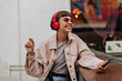 © Look! - Joyful teen girl with brunette hair listening to music outside. Short-haired lady in red headphones, pink sunglasses and light outfit smiles in cafe..