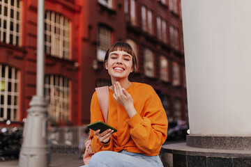 Young short-haired woman in orange sweatshirt smiles sincerely outside. Happy cool girl holds phone and sits outdoors.