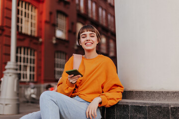 Wall Mural - Excited teen girl in orange sweatshirt leans on building and holds smartphone. Joyful woman in stylish hipster outfit smiles outside.