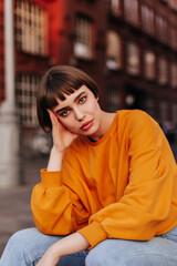 Wall Mural - Thoughtful brunette short-haired woman sits outside. Attractive young girl in orange sweatshirt and jeans looks into camera.