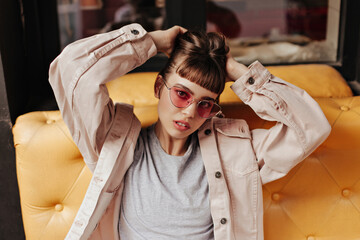 Wall Mural - Brunette woman in pink glasses sitting on yellow couch in cafe. Trendy girl with short hair in light jacket posing inside..