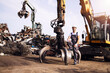 Portrait of junkyard worker with hardhat standing next to hydraulic industrial machine with claw attachment used for lifting scrap metal parts.