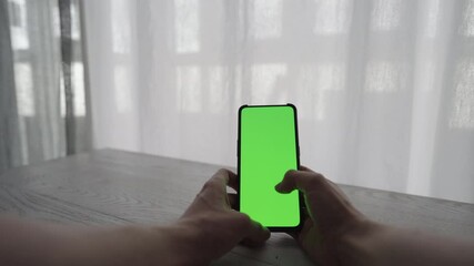 Wall Mural - Man showing and using smarphone with green screen while sitting behind white oak table