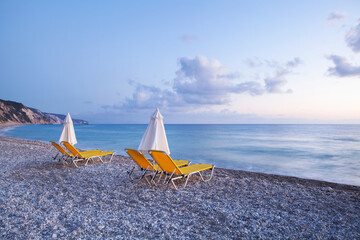 Holiday time, vacation, Panorama of the pebble coast with white pebbles with two yellow deck chairs and calm azure blue sea at dusk