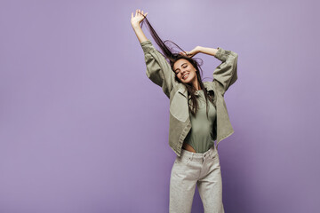 Wall Mural - Joyful girl with long brunette hair in cool t-shirt, modern olive jacket and wide pants smiling and posing with closed eyes.