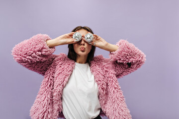 Wall Mural - Stylish brunette haired lady in plain t-shirt and long sleeve pink trendy sweater holding two small disco balls on lilac backdrop.