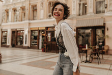 Optimistic Woman With Curly Hair In Jeans Smiling Sincerely In City. Cool Lady In Light Blouse With Black Lace Looking Into Camera At Street..