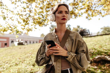 Wall Mural - Wonderful lady with short hair in white headphones and olive jacket looking away outdoors. Girl with handbag holds phone outside..