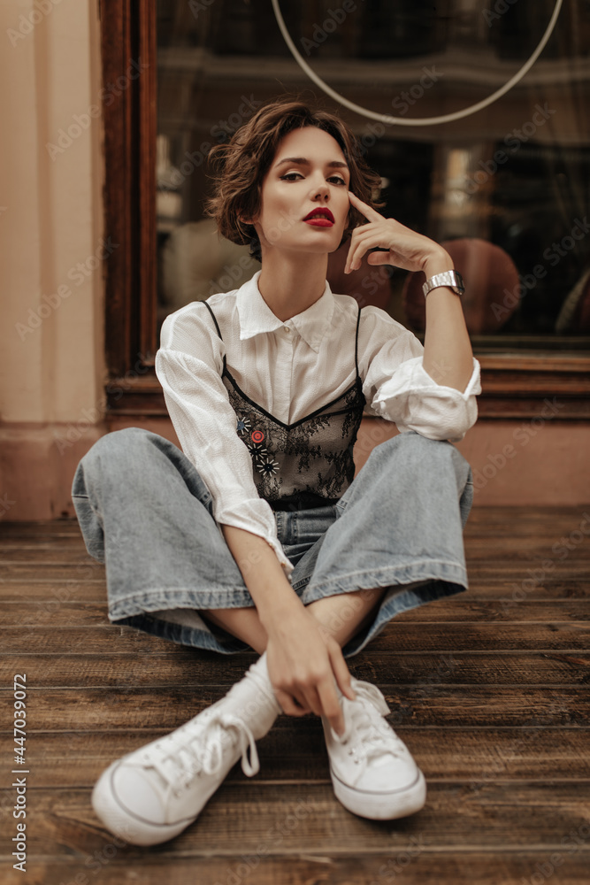 Stylish girl in jeans, white sneakers and blouse with lace sitting on floor at street. Modern woman with short hair poses outside.. - obrazy, fototapety, plakaty 