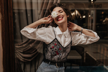 Wall Mural - Positive girl with short hair in long sleeve white and black shirt smiling sincerely in cafe. Cheerful lady in jeans holding flower inside..