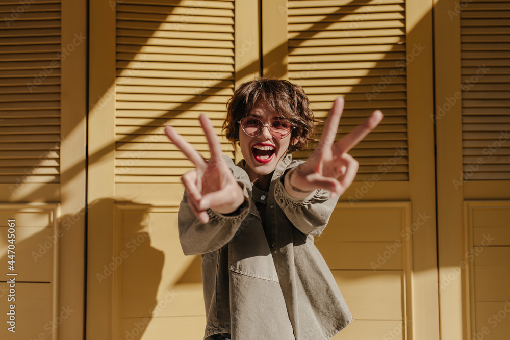 Good-humored woman with red lips showing peace sign on yellow background. Cool lady with curly hair in glasses smiles on bright backdrop.. - obrazy, fototapety, plakaty 