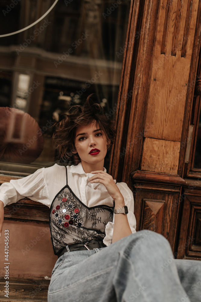 Fashionable lady in jeans with black lace and jeans poses on background of wooden door. Curly girl with bright red lips looks into camera at street.. - obrazy, fototapety, plakaty 