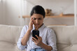Unhappy Indian millennial woman looking at phone screen, sitting on couch, reading bad news in message, upset young female holding smartphone, having problem with broken or discharged mobile device