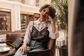 Wall Mural - Stylish lady with short hair in jeans and blouse looks into camera in restaurant. Happy girl with bright lipstick and wrist watch in cafe..